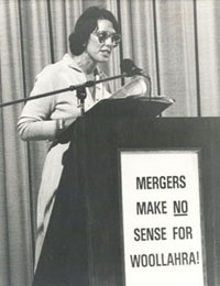 Rosemary speaking at a community meeting behind a lectern that says 'Mergers make no sense for Woollahra!